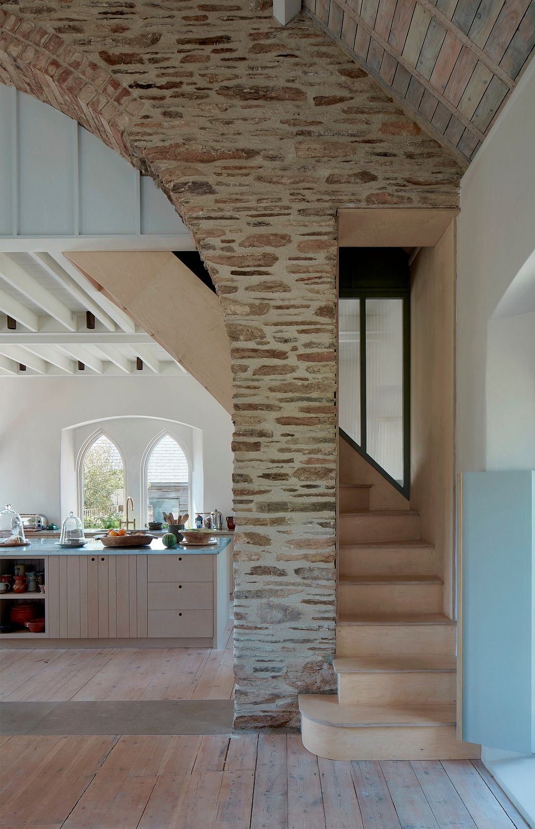 A modern kitchen with a rustic stone wall, wooden floors, and a curved staircase.