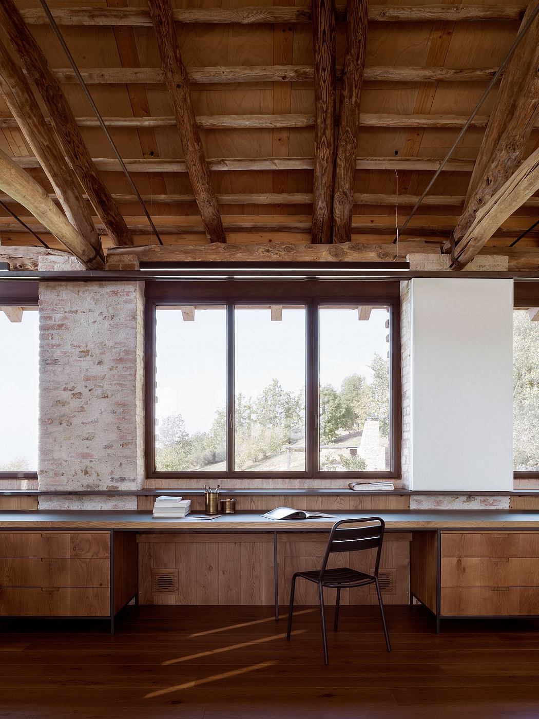 A cozy, rustic workspace with exposed wooden beams, large windows, and a simple metal chair.
