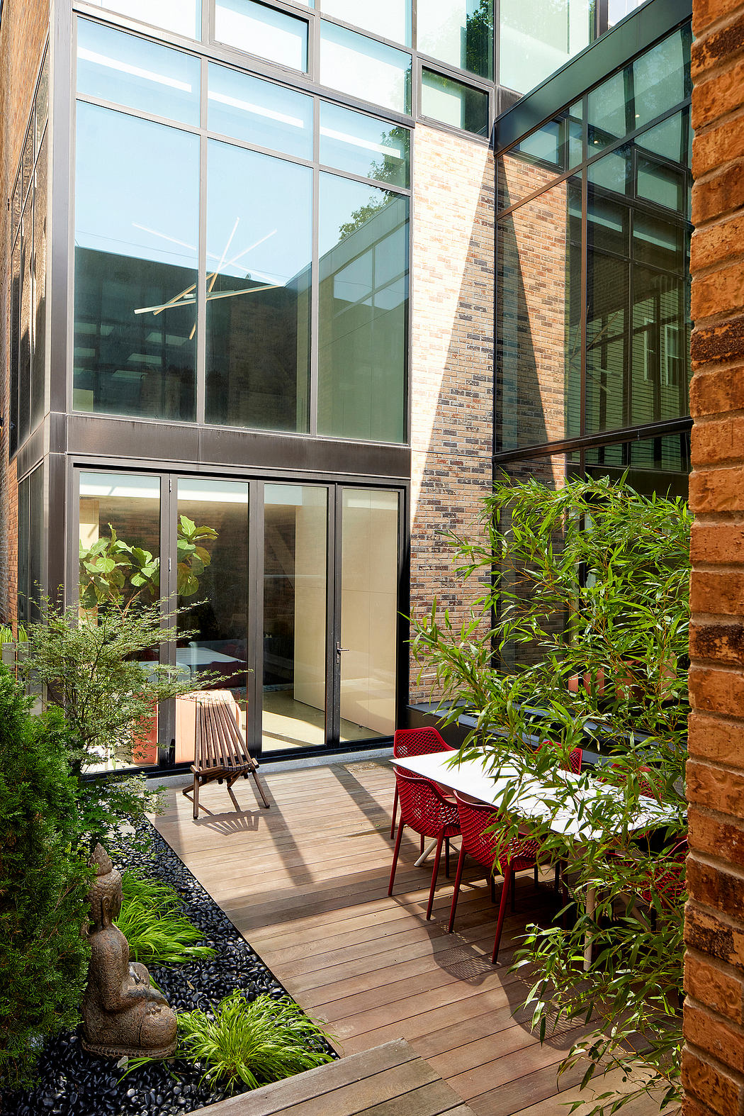 A modern glass-enclosed courtyard with a wooden deck, lush greenery, and red chairs.