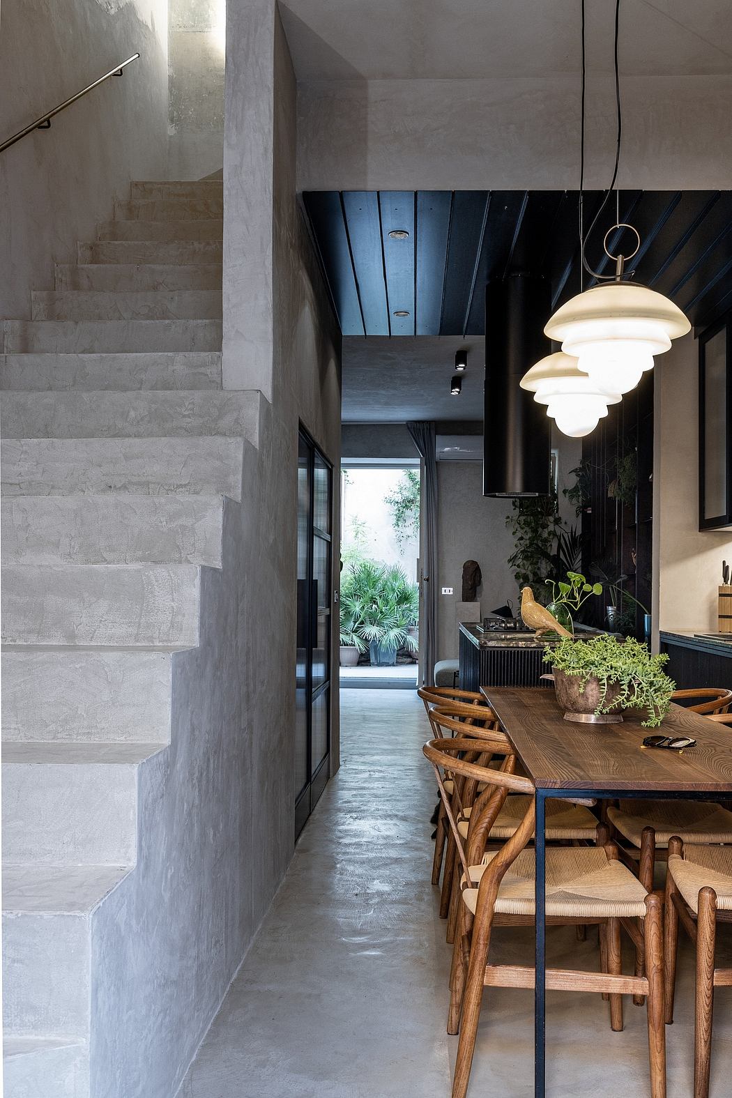 Minimalist concrete interior with wooden dining furniture, pendant lighting, and views of greenery.