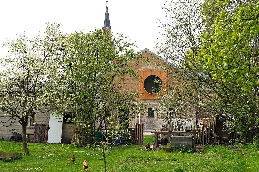 A quaint brick building with a distinctive steeple surrounded by lush greenery and chickens.