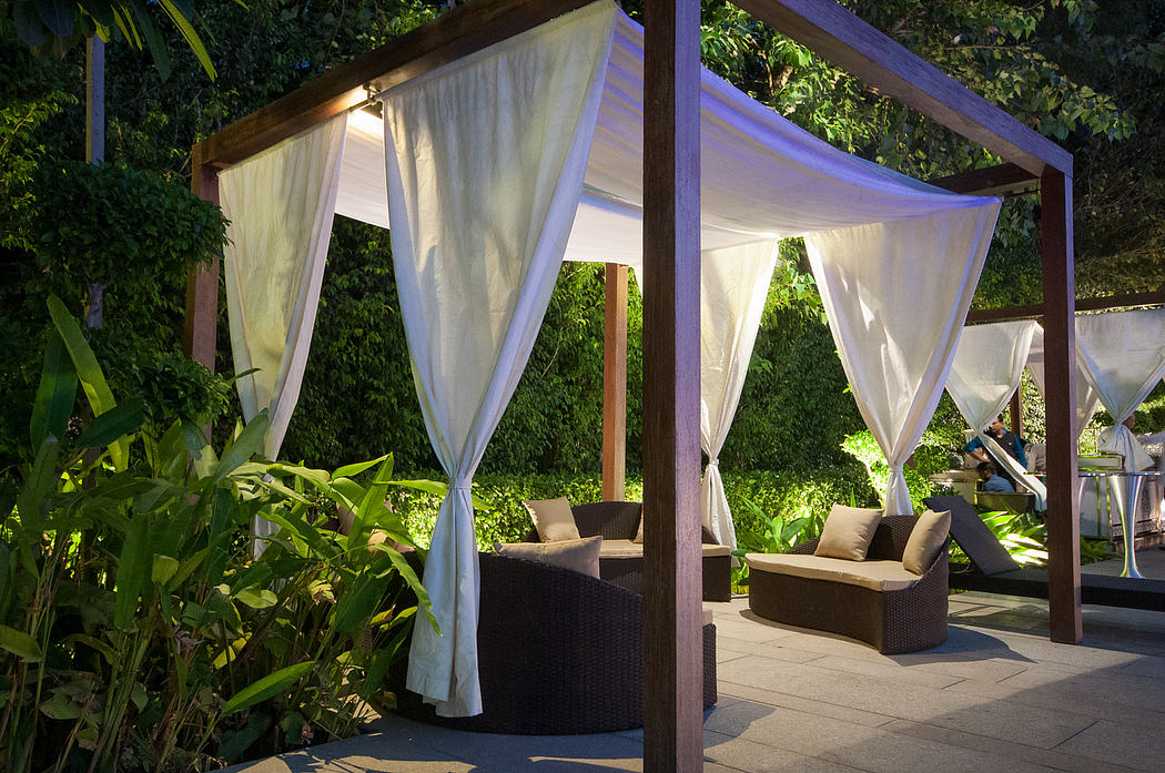 Cozy outdoor seating area with wooden canopy, white curtains, and lush greenery.