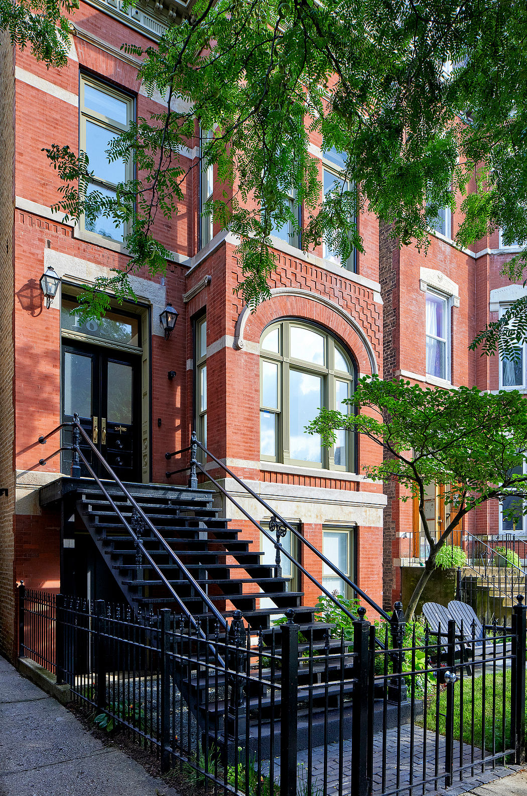 Ornate brick townhouse with arched window, wrought-iron fence, and lush greenery.