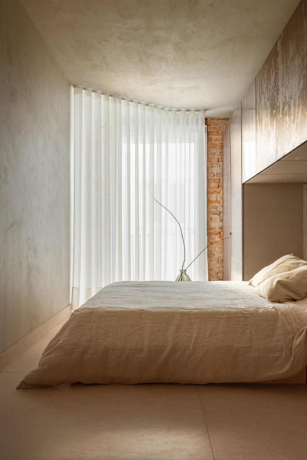 A minimalist bedroom with white curtains, a beige bed, and a brick accent wall.