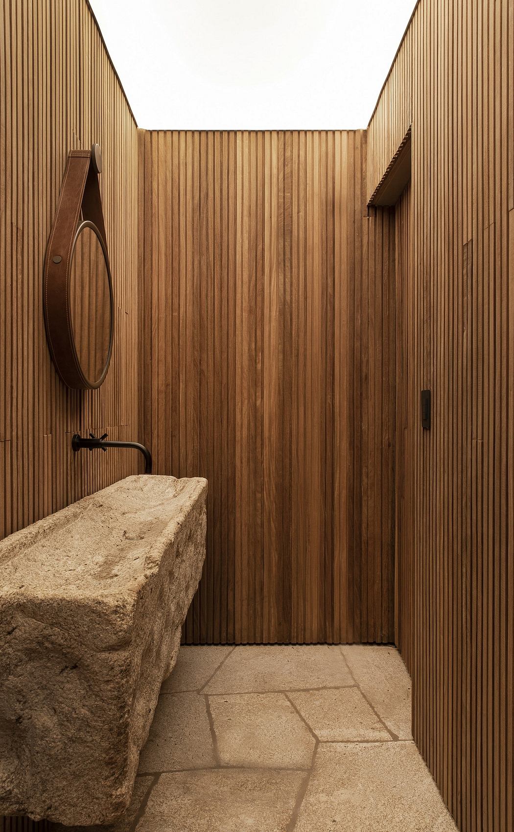 A minimalist bathroom with wooden paneled walls, a natural stone sink, and a round mirror.