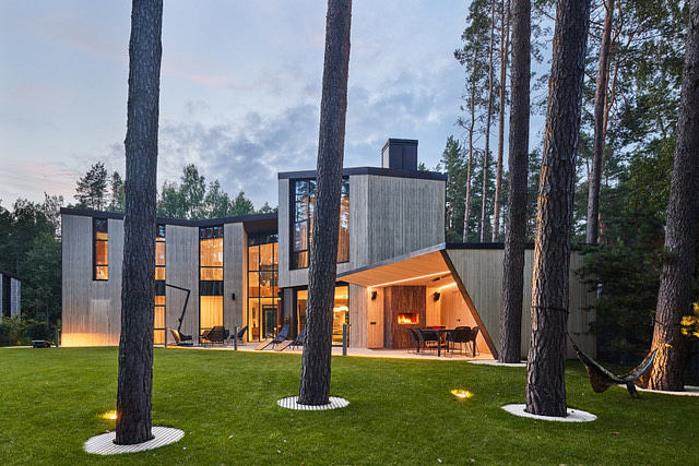 A modern, wooden house nestled in a pine forest, featuring large windows and patio.
