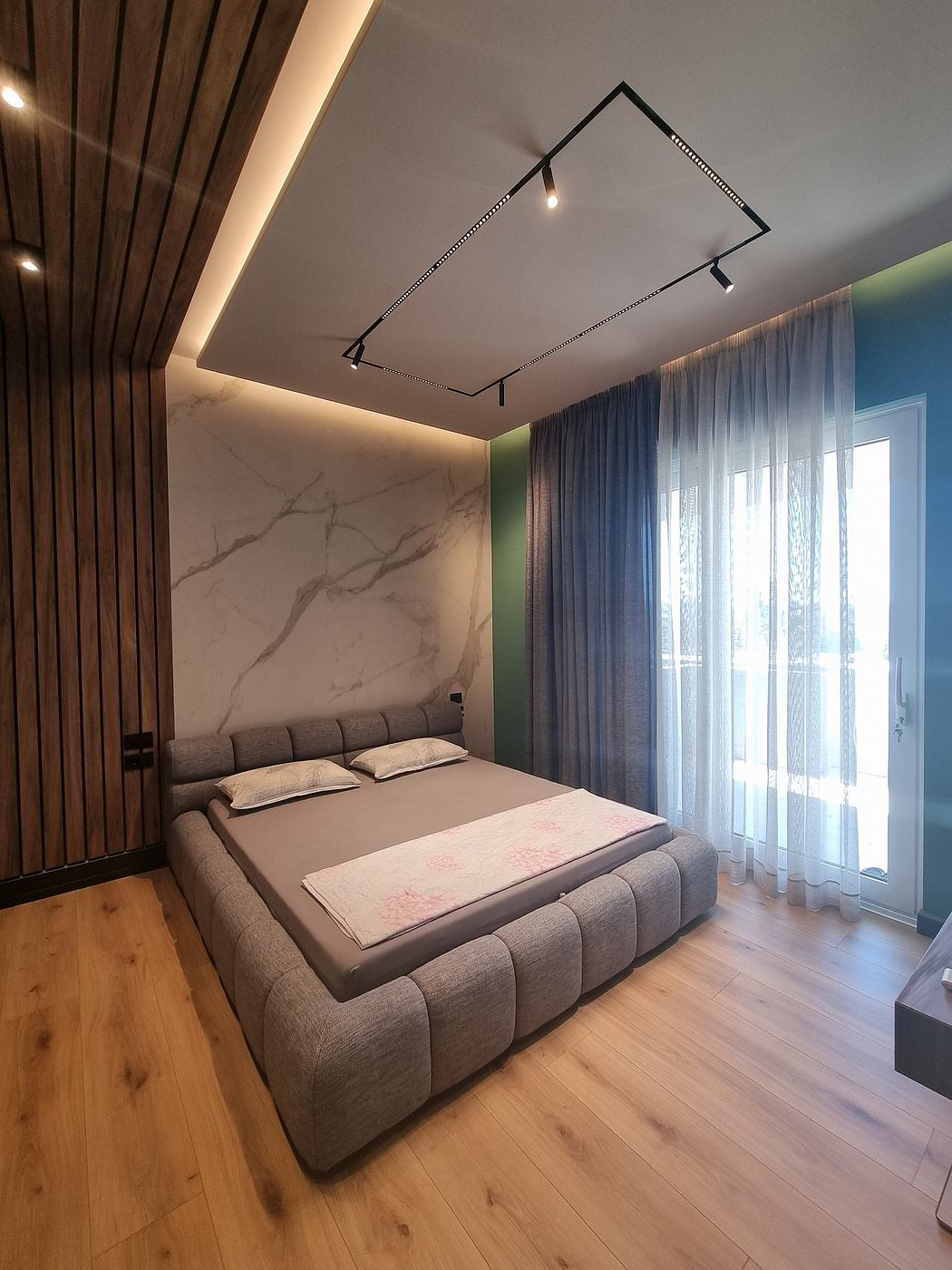 A modern bedroom with a marble accent wall, track lighting, and wood flooring. Stylish curtains frame the window.