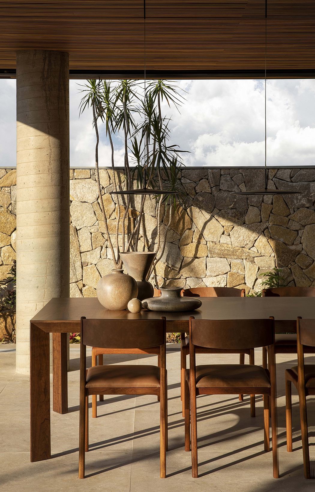 A modern, minimalist dining area with a stone wall, wooden furniture, and potted plants.