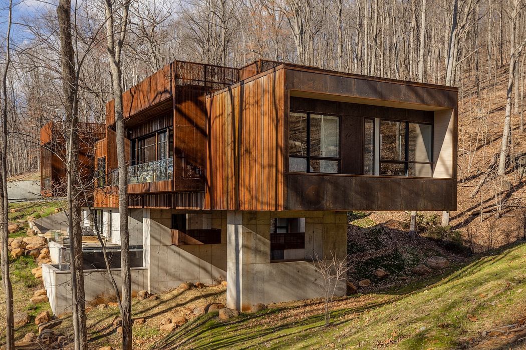 A modern, wooden cabin-style house with large windows and a cantilevered deck nestled in a forest.