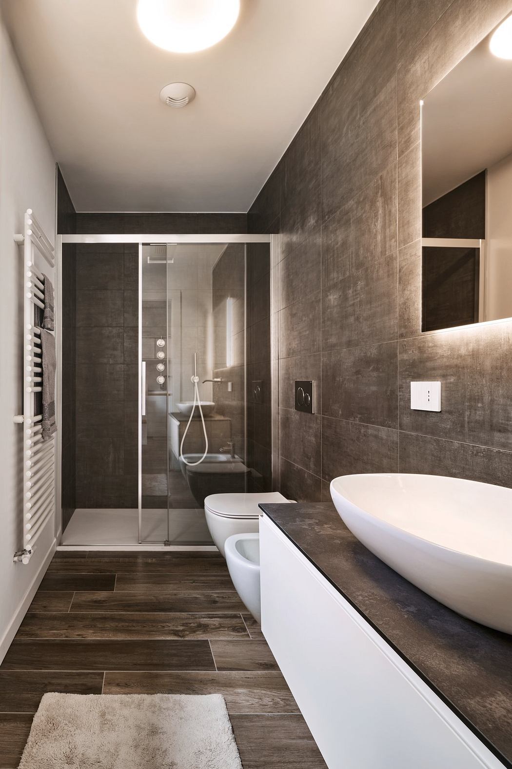 A modern bathroom with a sleek white bathtub, glass shower enclosure, and gray tiled walls.