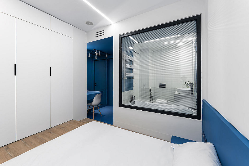 Modern, minimalist bedroom with white cabinets, blue accents, and large window overlooking bathroom.