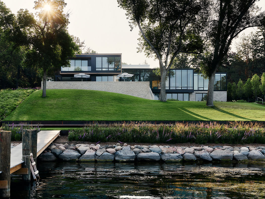 Sleek modern lakeside home with glass walls, lush greenery, and a wooden dock.