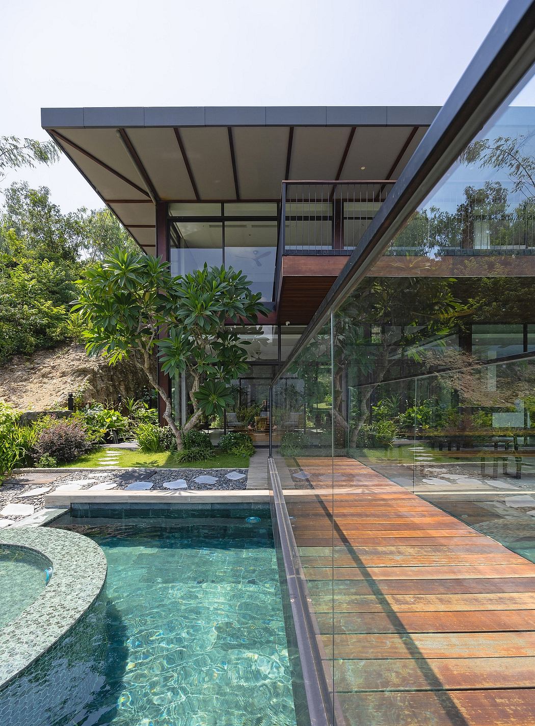 Sleek modern architecture with glass walls, wooden decking, and lush tropical landscaping.