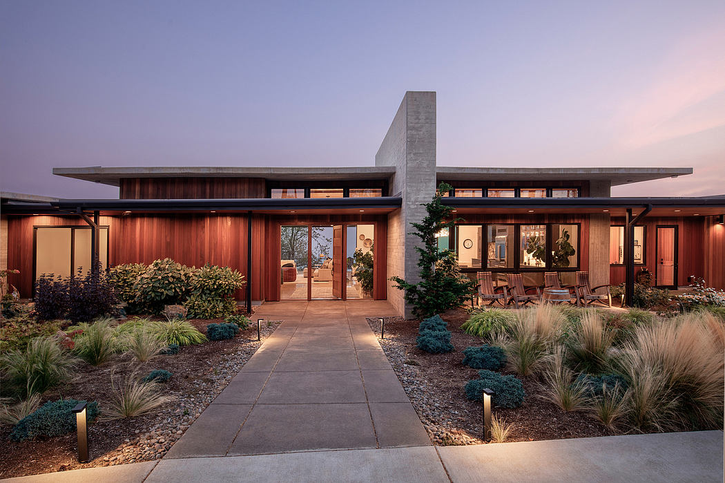 A modern, wood-paneled home with an expansive patio, concrete walkway, and landscaped grounds.