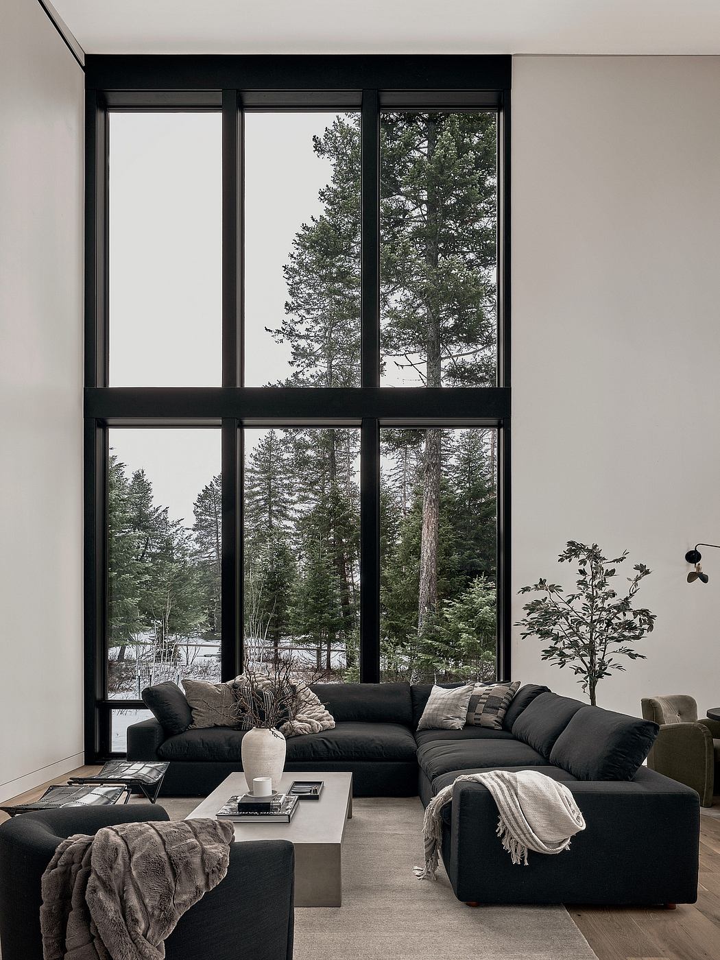 A modern, minimalist living room with large black-framed windows showcasing a serene forest view.