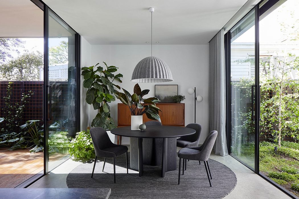 A modern, minimalist dining area with a circular wooden table, dark chairs, and lush plants.