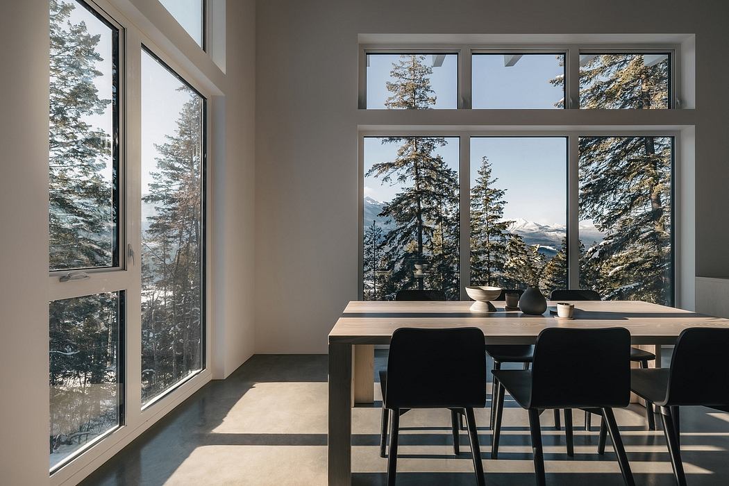Expansive windows showcase a snowy forest landscape; modern dining space with wood table.