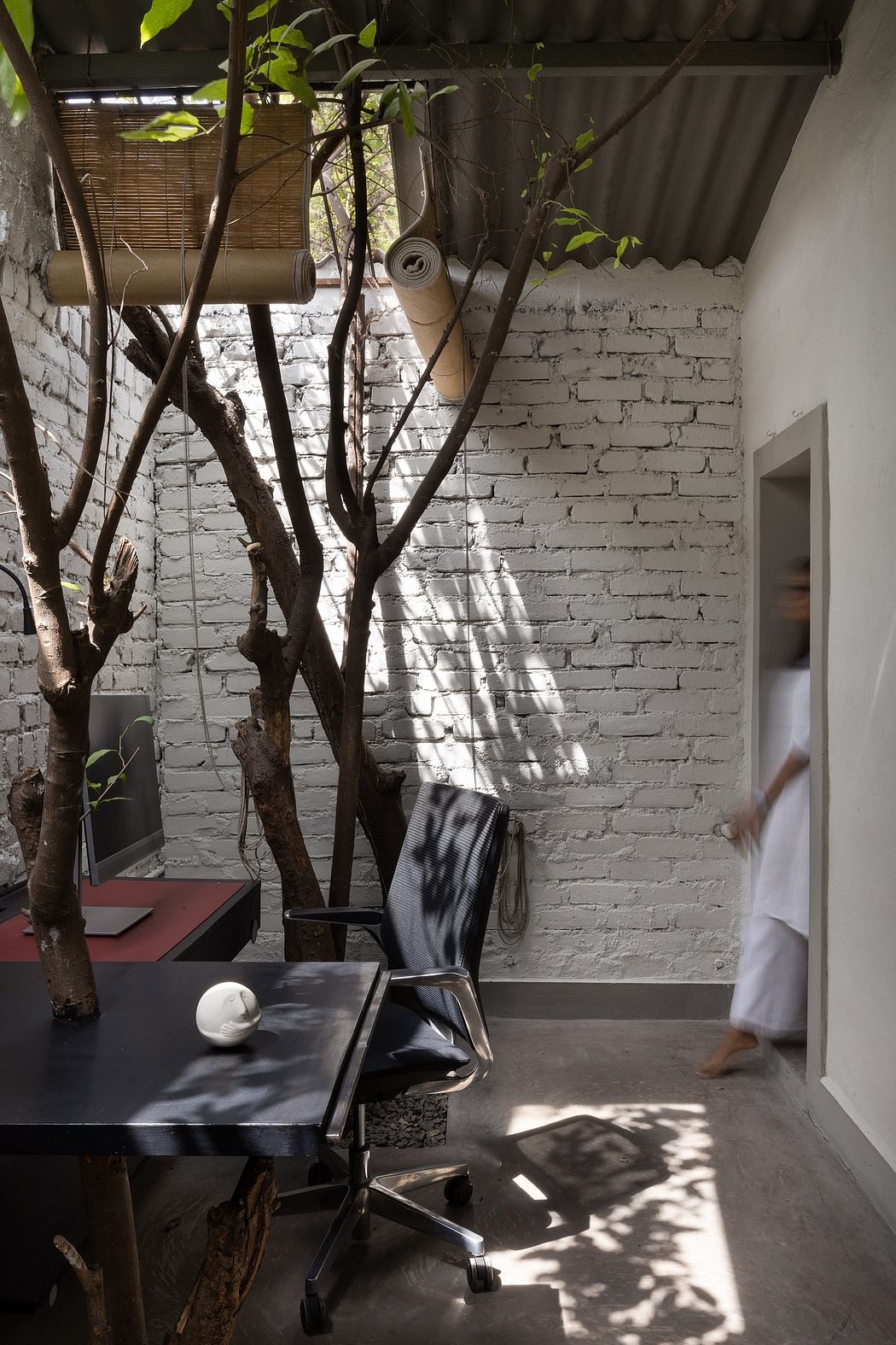 A workspace with a black desk, rolling chair, and leafy branches casting shadows on the brick wall.