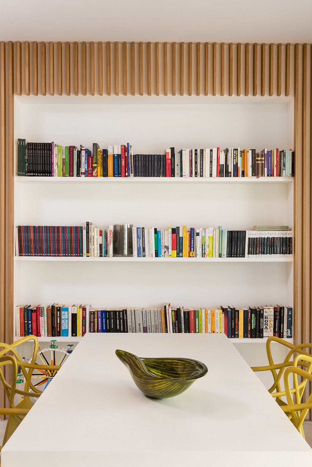 Minimalist white shelving with diverse book collection, wooden slats, and bright yellow chairs.