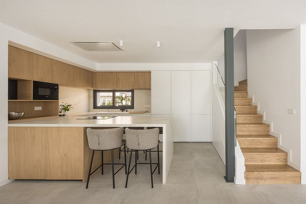 Minimalist kitchen with wood cabinetry, gray bar stools, and concrete flooring leading to stairs.