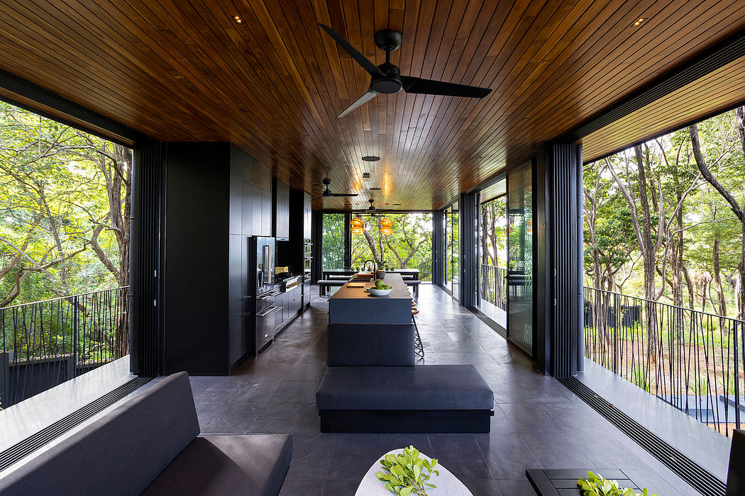 A modern, open-plan interior with a wooden ceiling, black kitchen island, and expansive windows overlooking nature.