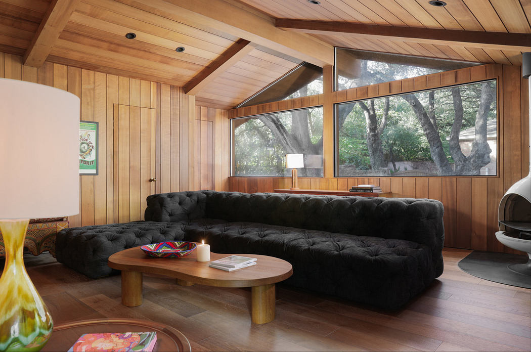 A cozy wooden cabin interior with a tufted black sofa, a low oval coffee table, and expansive windows overlooking the trees outside.