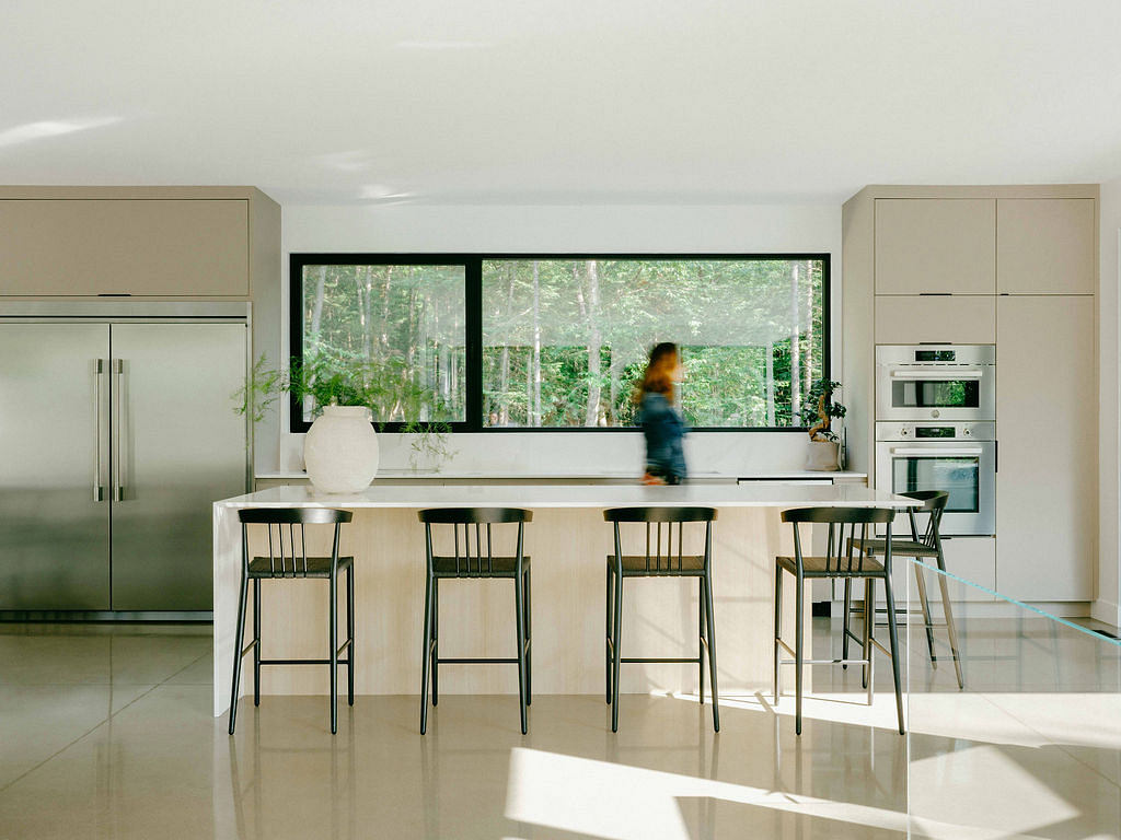 Spacious, modern kitchen with large windows overlooking lush forest scenery, sleek cabinetry, and bar seating.