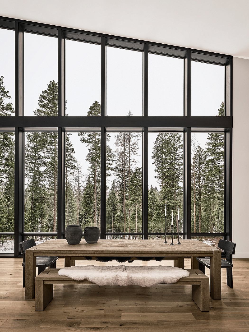 Spacious dining area with wooden table and bench, framed by floor-to-ceiling windows showcasing a scenic forest view.