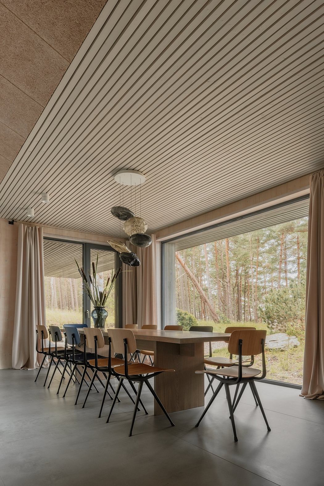 A spacious, modern dining area with a long wooden table, metal chairs, and a large window overlooking a forested landscape.