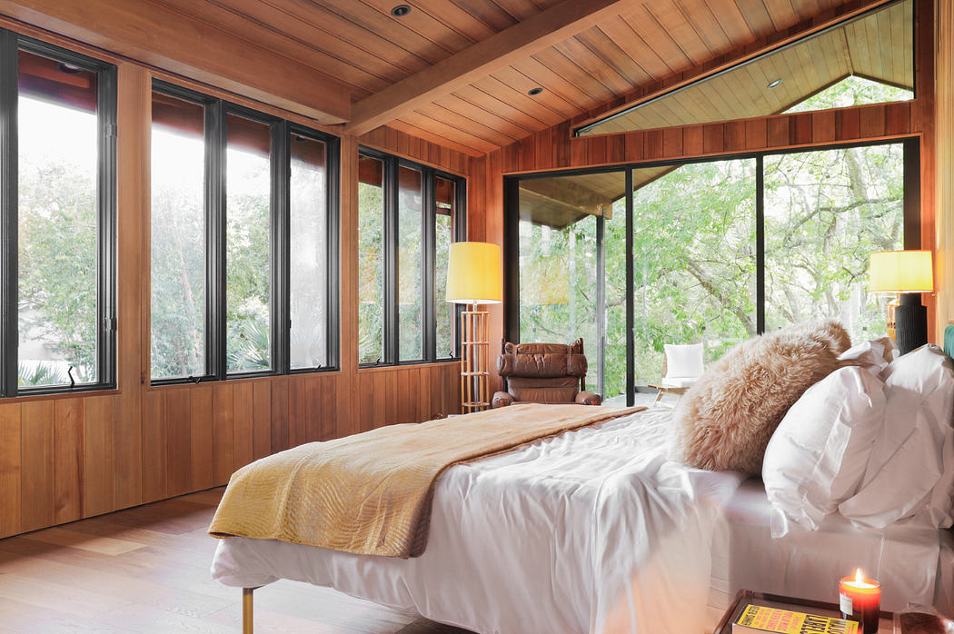 A cozy bedroom with a wooden ceiling, large windows, and natural lighting, showcasing a rustic design.