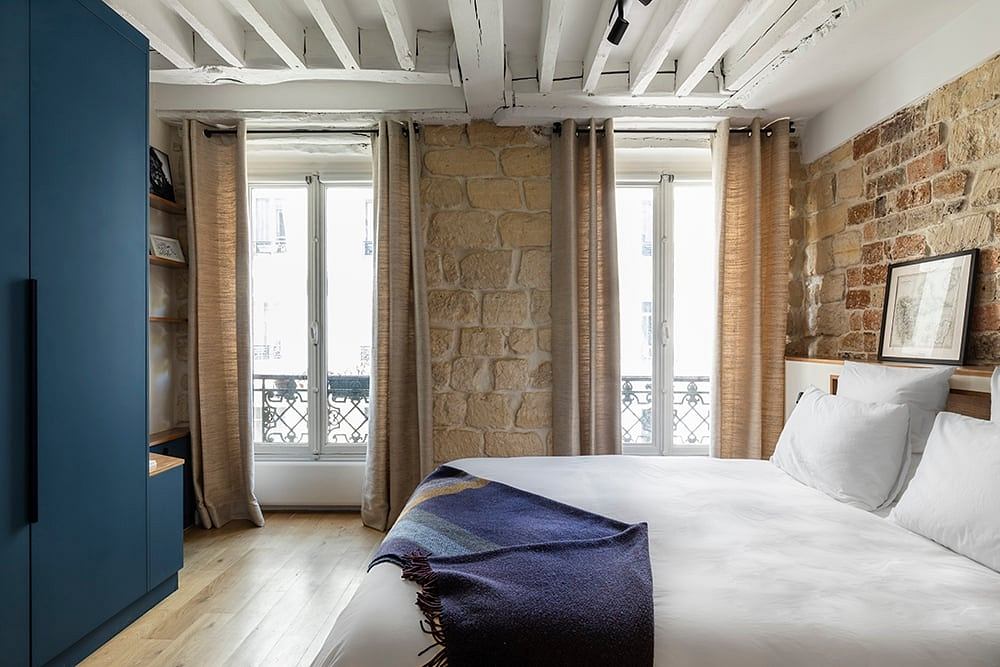 Rustic bedroom with exposed stone walls, high ceilings, and large windows overlooking balcony.