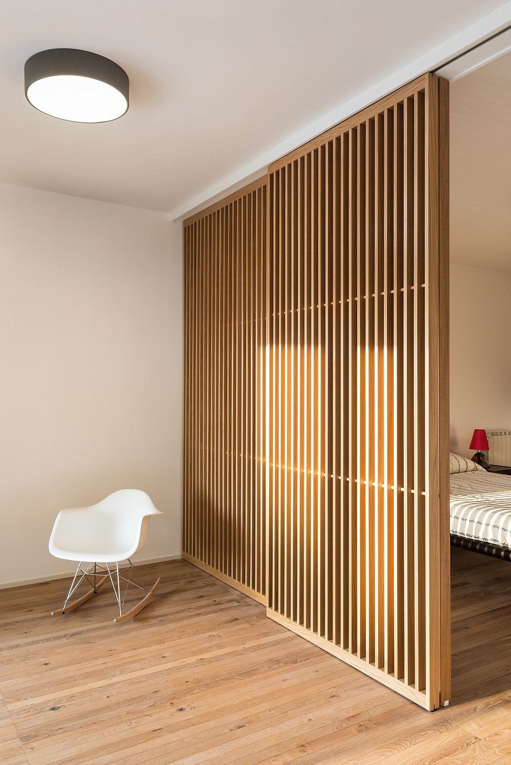 Minimalist bedroom with wooden slat wall, hardwood floor, and Eames-style rocking chair.