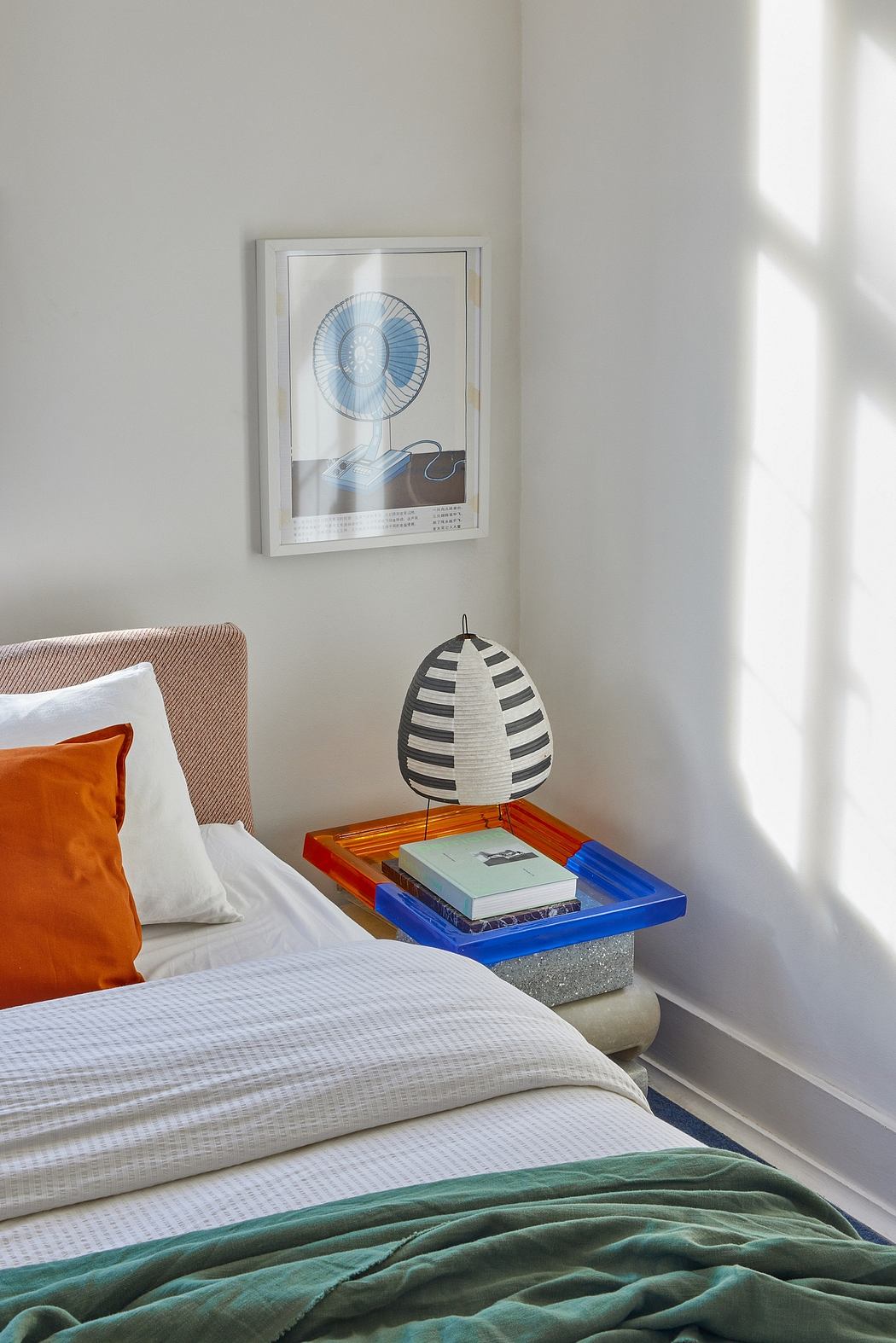 A cozy bedroom with a framed fan artwork, books, and a patterned lamp on a colorful nightstand.