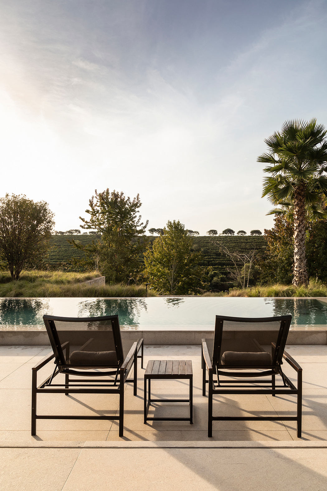 Two black lounge chairs and a wooden table by a pool, with a lush landscape in the background.