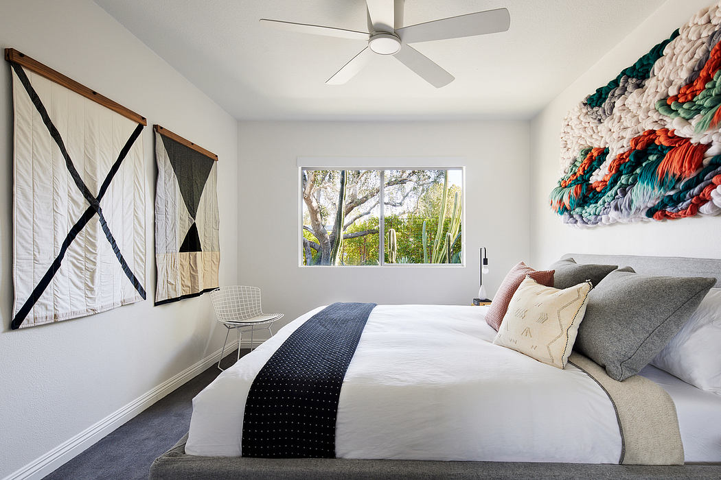 A modern bedroom with a large window, minimalist artwork, and a colorful textile wall hanging.