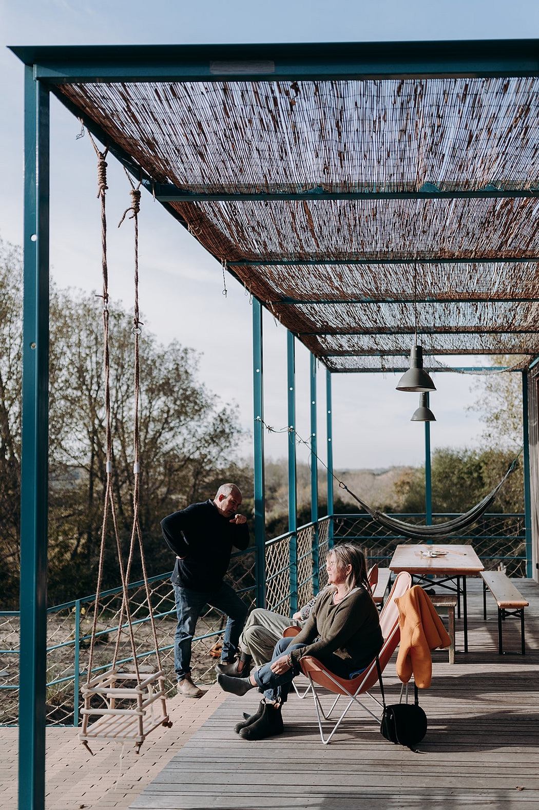 Covered patio with rustic thatched roof, metal lanterns, and chairs on wooden deck overlooking nature.