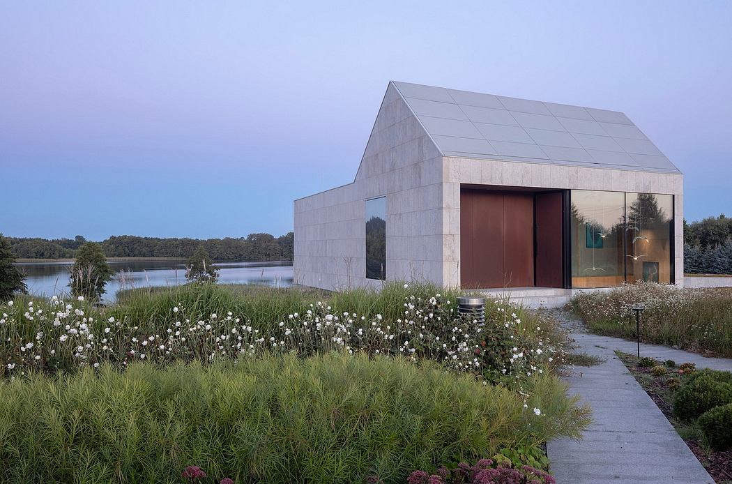 A modern concrete building with a slanted roof and large wooden doors stands in a grassy field near a lake.