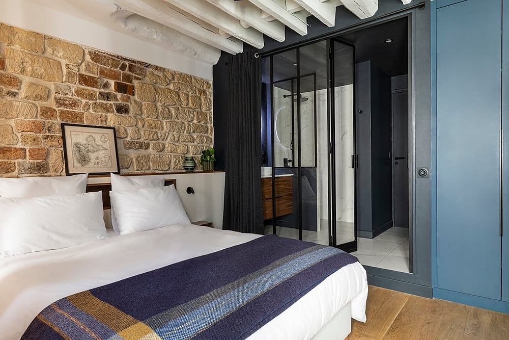 A cozy bedroom with exposed brick walls, wooden beams, and a modern, black-framed glass wall.
