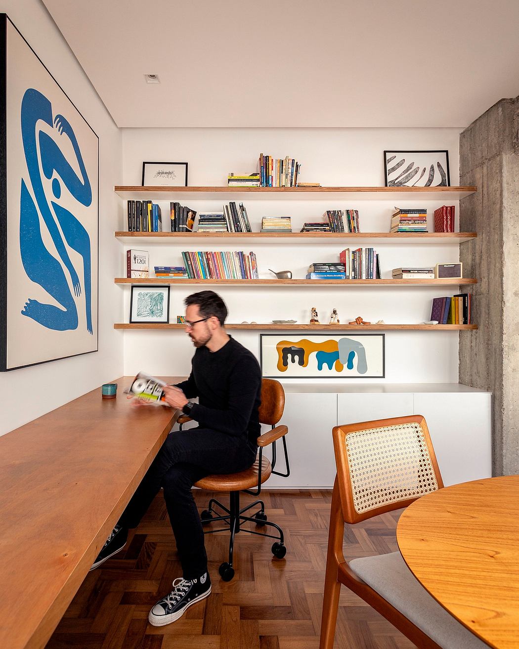 A cozy home workspace with wooden shelves, artwork, and a person seated at a wooden desk.