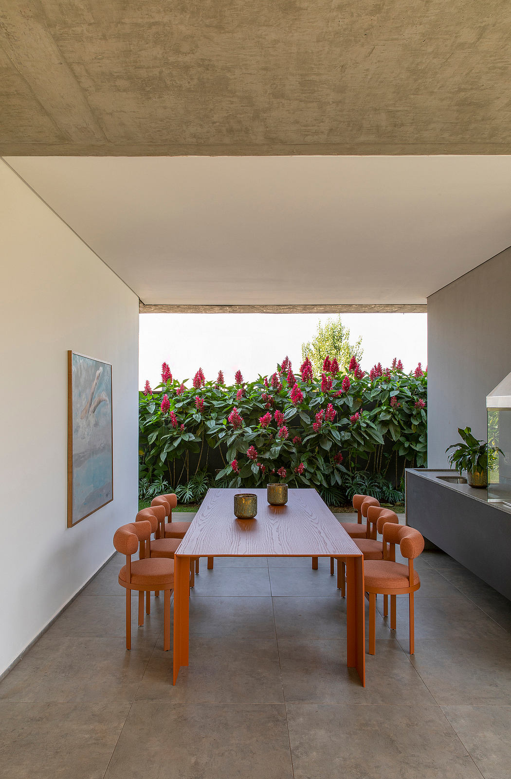 A modern dining area with a wooden table and chairs, flanked by vibrant foliage through a glass wall.