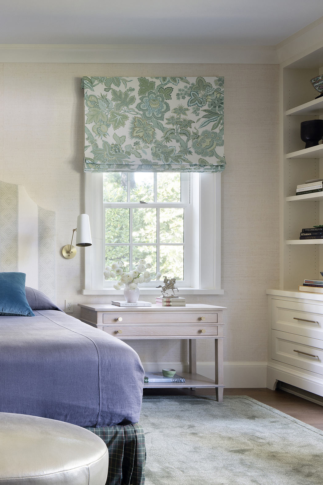 A cozy bedroom with a floral roman blind, white furniture, and blue bedding.