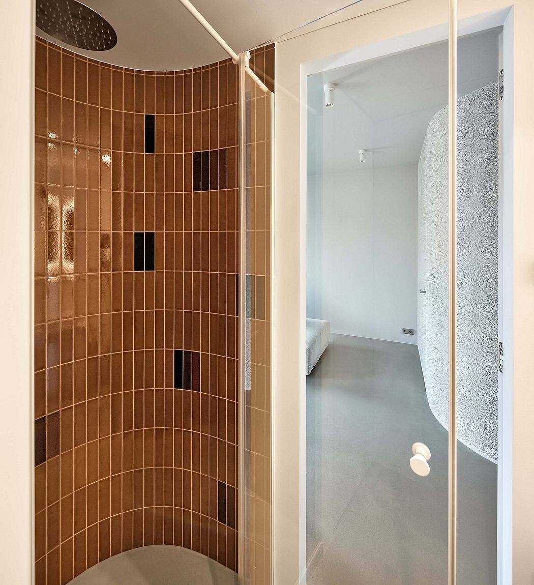 Curved shower tiled in warm brown hues, leading to a minimalist bedroom space.