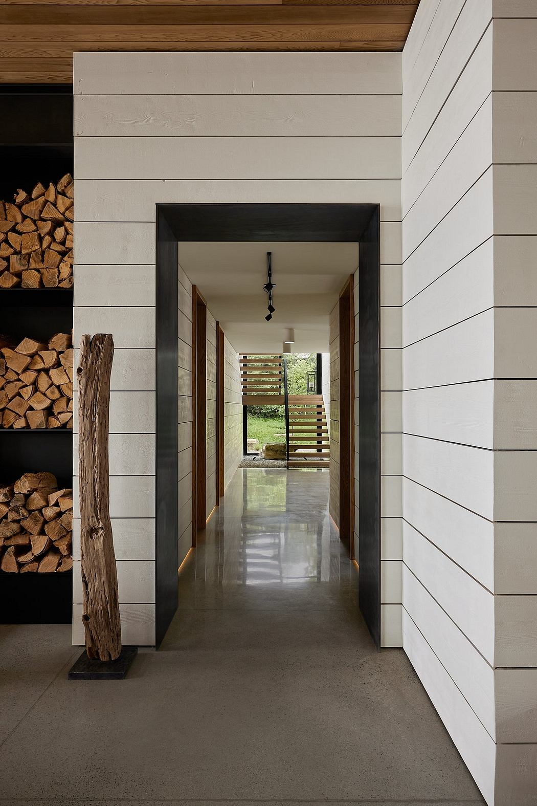 A modern architectural hallway featuring wooden beams, white paneled walls, and a glass door leading to the outdoors.