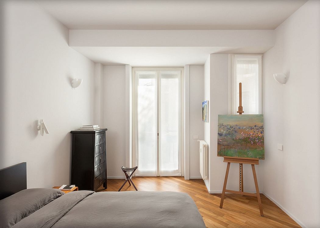 Bright, minimalist bedroom with large windows, wooden floors, and an easel displaying artwork.