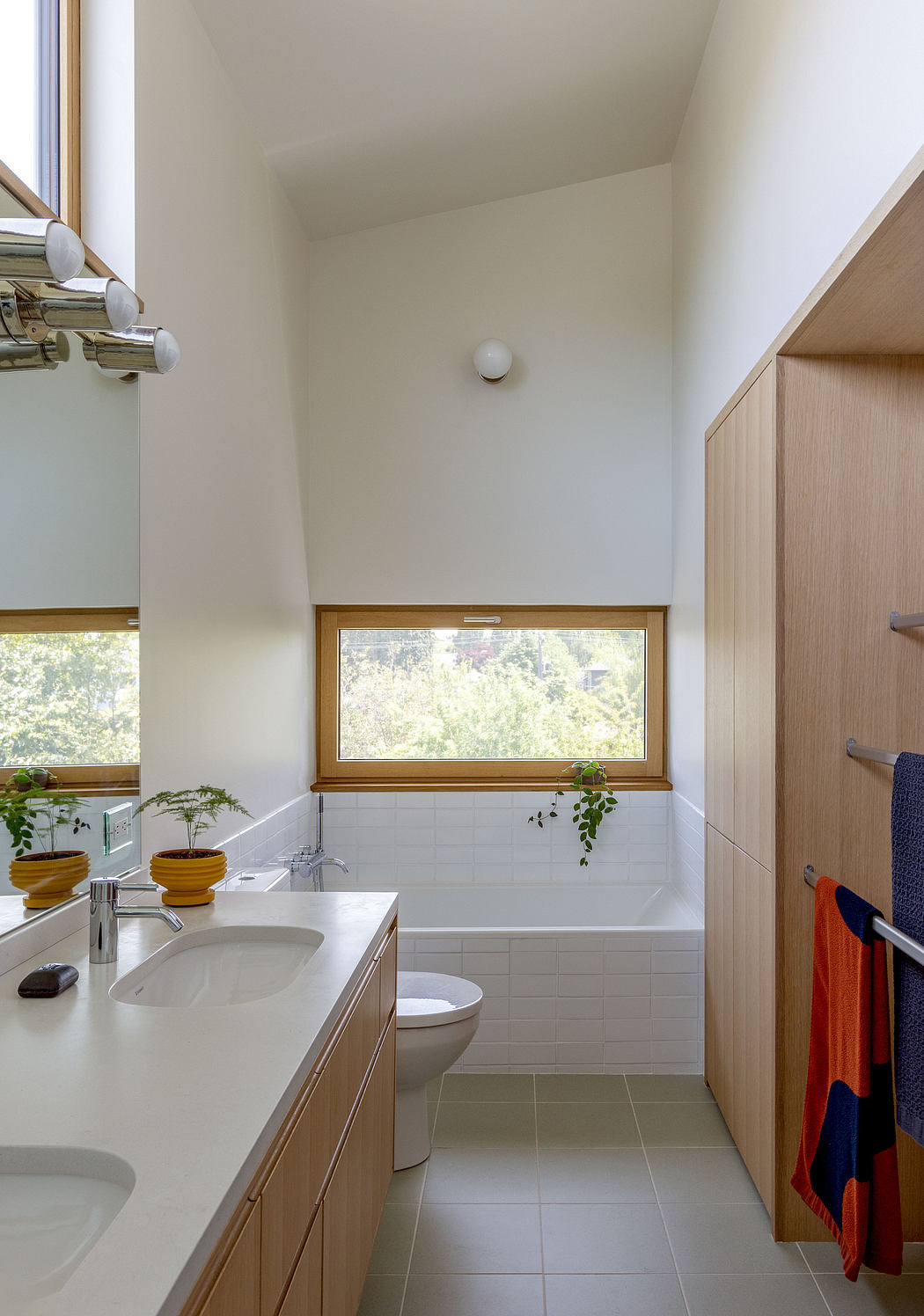 A modern bathroom with white walls, wooden cabinetry, and a large window overlooking greenery.