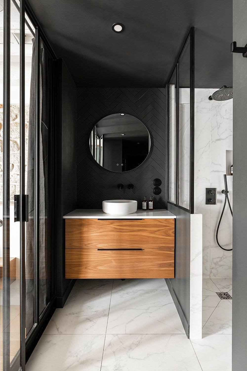 Sleek, modern bathroom with herringbone-patterned black tiles, marble vanity, and round mirror.