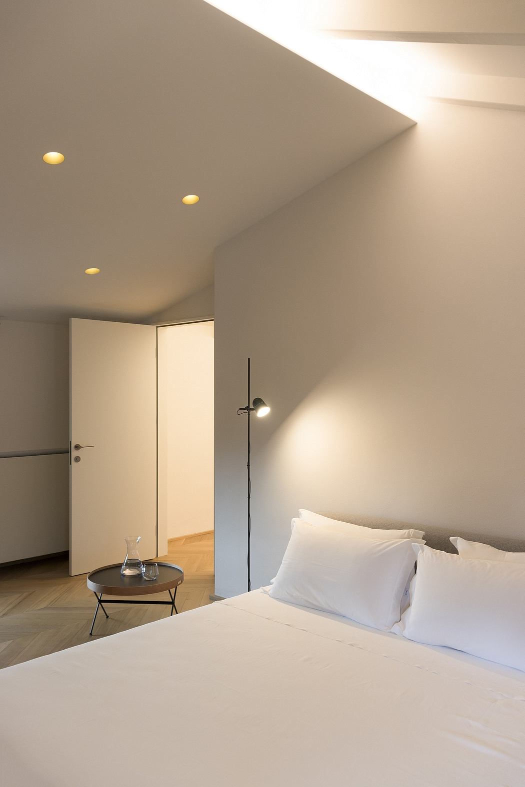 Cozy minimalist bedroom with recessed ceiling lighting, white bedding, and a dark side table.