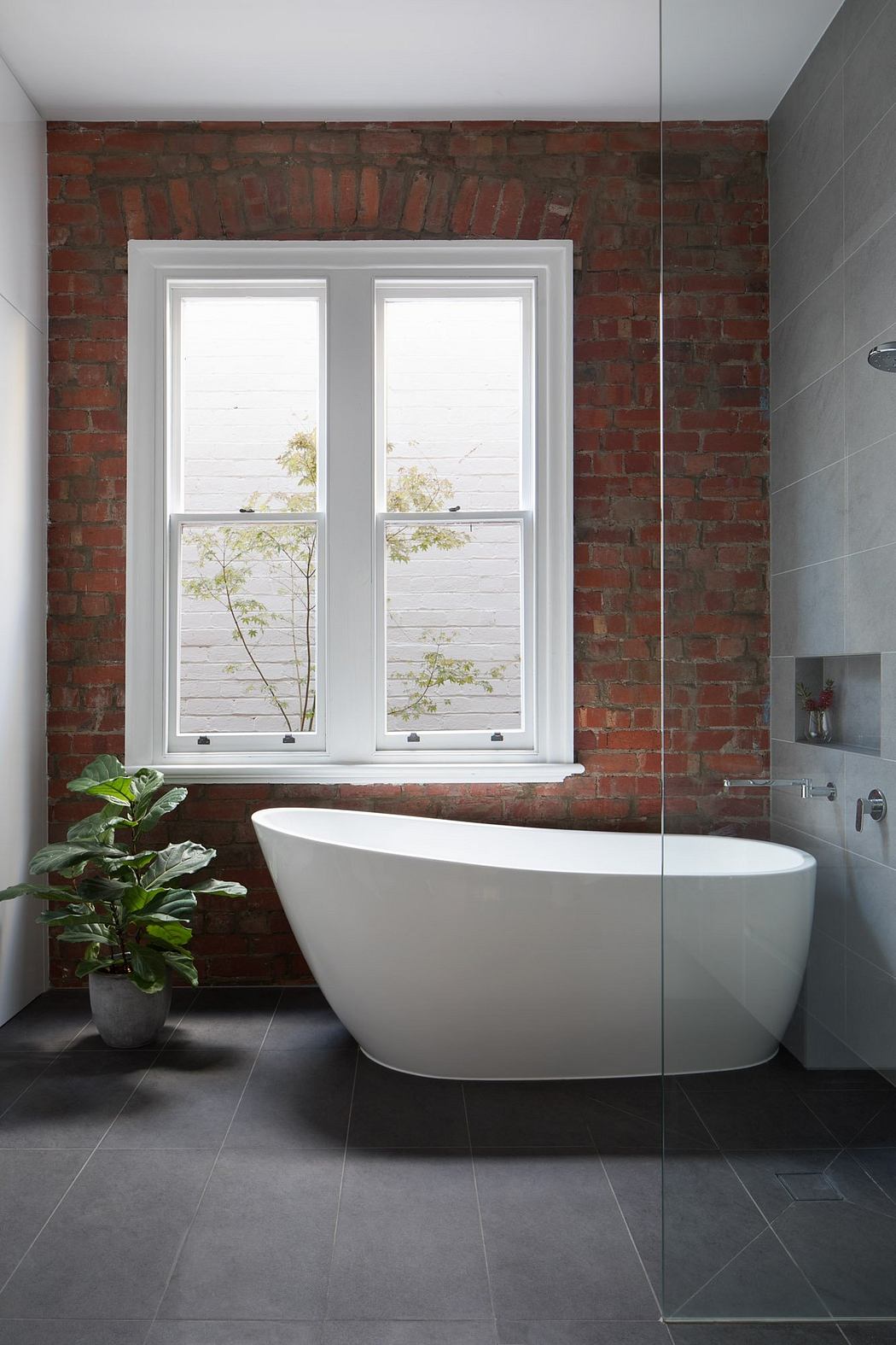 Modern bathroom with exposed brick walls, freestanding tub, and large windows overlooking nature.