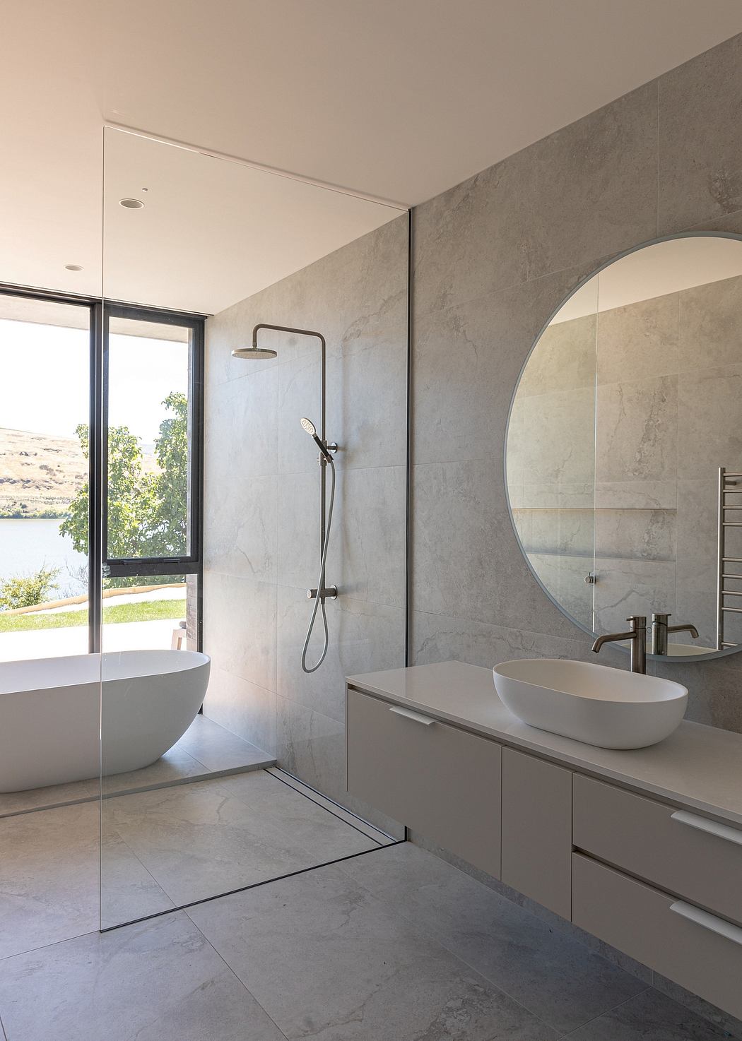 Spacious modern bathroom with sleek grey tiles, freestanding tub, and circular mirror.