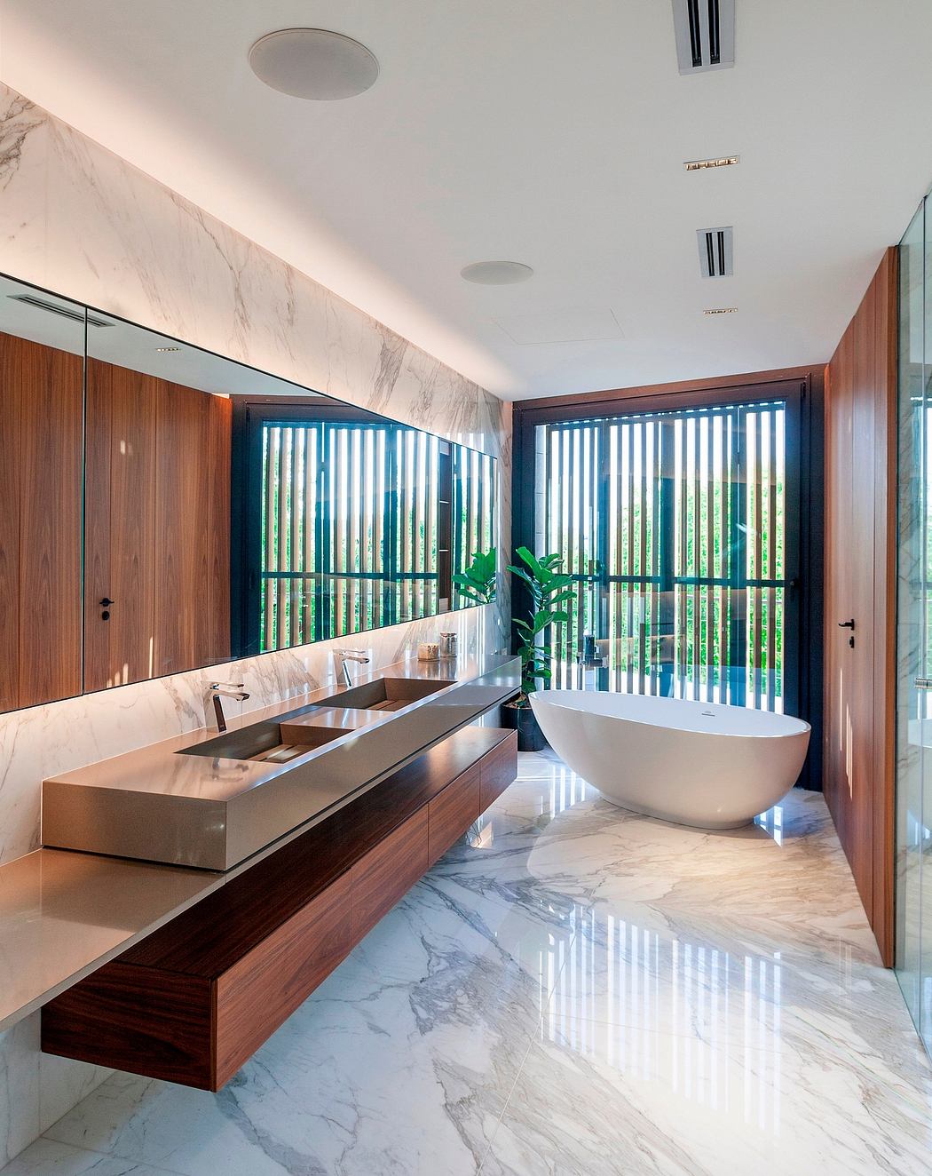 Luxurious bath with sleek wooden vanity, marble walls, and freestanding tub overlooking outdoor greenery.